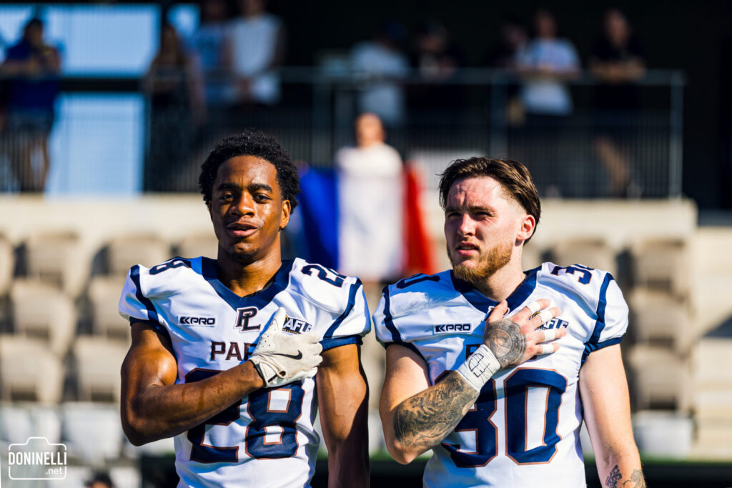Two American football players in white jerseys, numbers 28 and 30, stand with hands over their hearts, a French flag in the background.